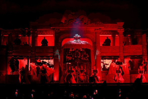 Lady Gaga in blood red Victorian-era dress. Her stage is lit up in red lights, evoking the feeling of hell. Underneath her dress' gigantic skirt is a cage holding people captive while they squirm and dance in pain.