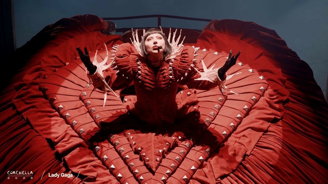 Lady Gaga in blood red Victorian-era dress. She looks up to the spotlight shining down upon her.
