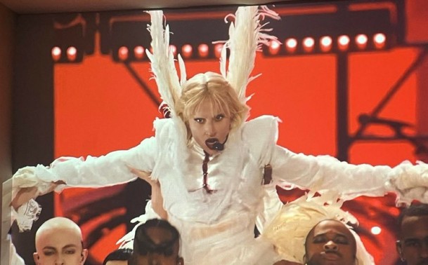 Lady Gaga wearing a white outfit with huge wing-like structures on the back of her head. Her dress has blood red stiches around the chest.