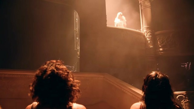 Lady Gaga shining in bright warm lights as she looks on freom a balcony held by corinthian pillars. People downstairs look on to her.