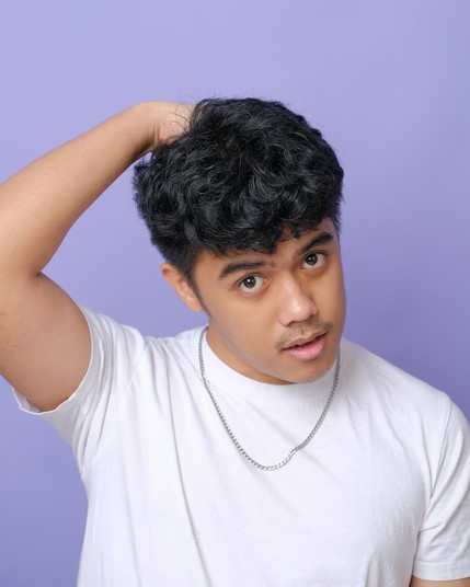 A boy in his mid-20s in a white shirt and a simple metal chain for a necklace. He puts one of his hands on the back of his head. He looks straight into the camera as if to beg for you to like him. The lavender background gives him a soft aura, but what really boils underneath this tender façade?