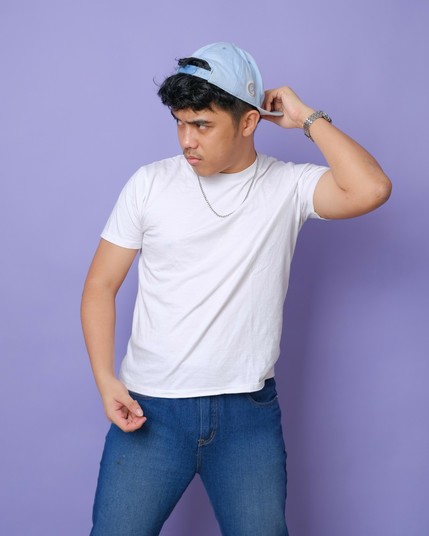 A boy in his mid-20s wearing a white shirt and blue jeans. He wears a light blue cap backwards, holding its brim with one of his hands as he looks to the side. Is he embarrassed, or merely playing coy?