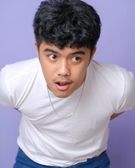 A boy in his mid-20s in a white shirt, blue jeans, and a metal chain necklace. His eyes are wide as he looks to the side, mouth agape. Time has caught up to him, and he is supposed to have become a man now. What happened?