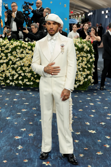 Lewis Hamilton wearing an all-cream ensemble. He wears a beret, a tuxedo, a satin bow around the collar of his white undershirt, a pair of trousers, and glossy black shoes.