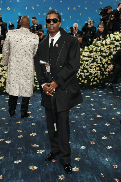 Asap Rocky in an oversized black coat on top of a black vest that covers a white undershirt with black tie. His collar is embellished with a fractal-style necklace of silver and diambonds. He holds a black umbrella whose handle is styled to look similar to a gun and the rest of the umbrella's stem is studded with crystals.