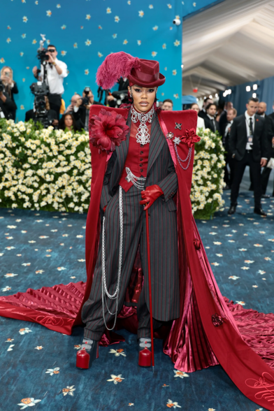 Teyana Taylor is serving peak dandy glam in a custom red and charcoal pinstripe ensemble that fuses masculine tailoring with high-drama couture — complete with a sharp-shouldered cape, layered chains, corseted waistcoat, and a feather-topped fedora. The look is a full theatrical fantasy, dripping in textures from velvet to satin, finished off with leather gloves, chunky platforms, and unapologetic attitude.