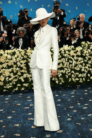 Zendaya wearing a wide-brimmed white hat, a cinched tuxedo on top of a white shirt and white satin tie. 