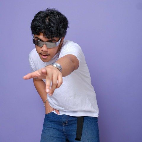 A dude in his mid-20s wearing silver sunglasses. His right hand is in his pocket, while his left hand points to the camera as he wears a smug look on his face.