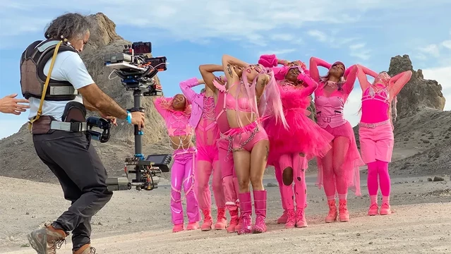 Lady Gaga and her dance team in bright pink in the desert. A camera man is shown holding a bunch of videography equipment, with an iPhone attached to it instead of a profesisonal camera.