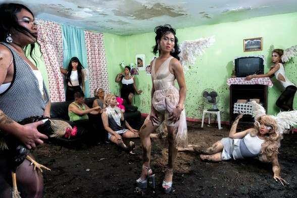 Sassa Gurl stands at the front of a crowded room with green walls and a damaged ceiling, wearing a patterned dress and holding a cigarette. The muddy floor is strewn with scattered furniture, a television, a fan, and various people in costumes, including angel wings and high heels.