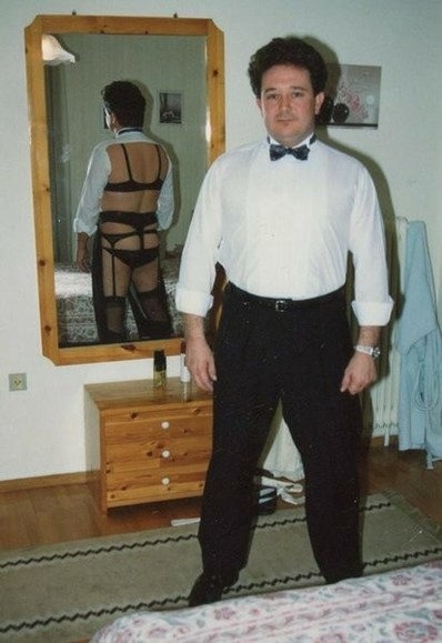 A man stands confidently in formal attire, while the mirror behind him reveals he's wearing lingerie beneath his suit. The contrast between his composed front and the unexpected reflection creates a humorous and subversive twist.

