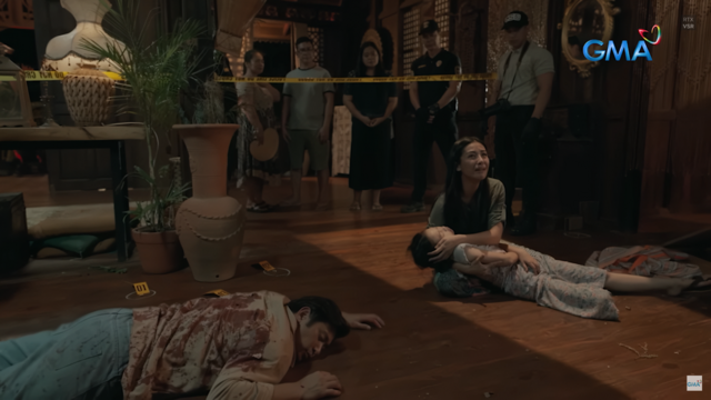 Danaya clutches her lifeless daughter Gaeia in anguish, surrounded by the aftermath of a brutal crime scene. Her husband Aquil lies motionless nearby, drenched in blood, as investigators and onlookers watch in stunned silence.
