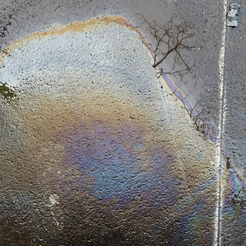 Das Bild zeigt ein Farbfoto, 1:1, das heißt quasi im CD-Format, eine Öl- beziehungsweise Benzin-Wasserpfütze mit blau-lila Zentrum und braun-gelbem Drumherum auf dunklem Asphalt, Bäume spiegeln sich, ein weißer Streifen rechts, ein Stück Alufolie.