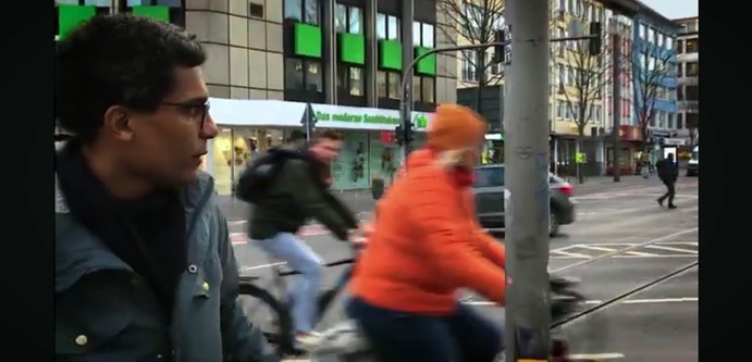 Screenshot eines Videos von Marvin Oppong, ein Mann im Profil, mit Brille, in Parka, Fahrradfahrerinnen, ein Laternenmast, im Hintergrund eine Straßenkreuzung, Geschäftsgebäude.