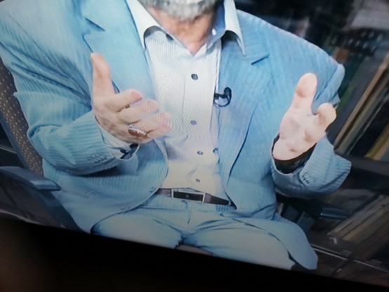 Bild: Farbfoto einer Interviewszene mit einem Intellektuellen der Revolutionsgarde in Teil 2 von _Irans heimliche Herrscher - Die Revolutionsgarden_, ein sitzender Mann, kein Gesicht, im hellblauen Anzug hält seine Hände nach vorne.
