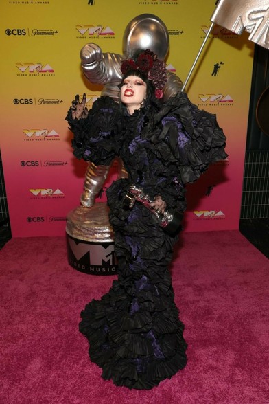 Lady Gaga, with her face painted pale and dramatic makeup, wears an elaborate purple and black ruffled gown with a large floral headpiece. She stands on a pink carpet holding an MTV VMA Moon Person trophy, next to a larger silver Moon Man statue.