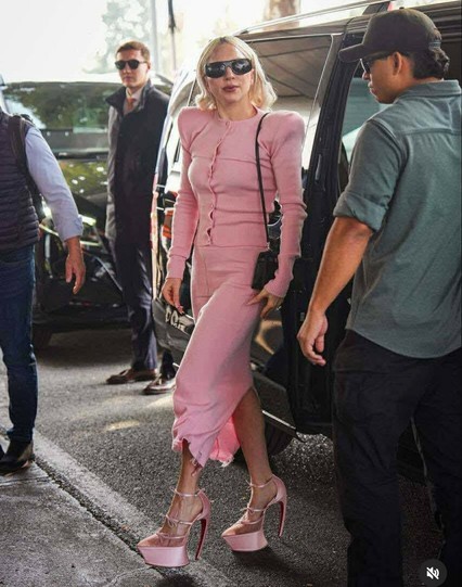 Lady Gaga in a monochromatic outfit in a soft, bubblegum pink hue, comprising a close-fitting, long-sleeved knit cardigan with padded, structured shoulders and a matching skirt that hits around mid-calf and features a raw, ruffled hemline. She completes the dramatic look with extraordinarily high-heeled platform shoes in the same pink, which feature a distinct, curved, stiletto-like heel that sweeps back without a visible arch.