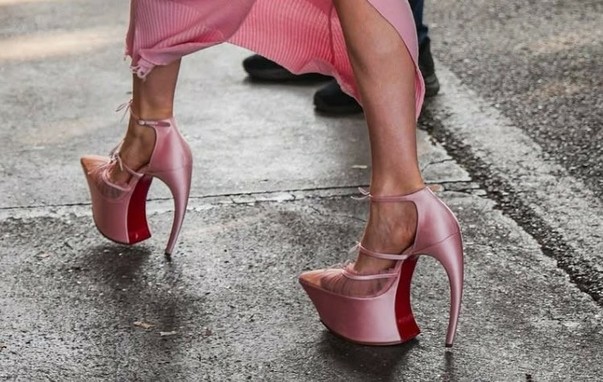 Lady Gaga's extremely high-heeled platform shoes with a unique, curved heel that sweeps dramatically backward without touching the ground under the arch. They are made of a pink satin or silk-like material, possibly with sheer mesh detailing over the toes, and feature ankle straps.