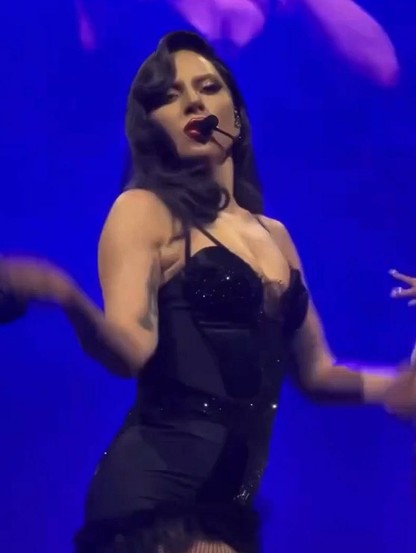 A striking image of Lady Gaga performing live on stage. She is wearing a black corset-style outfit and has dark hair styled in a vintage Hollywood wave. The background is lit with a dramatic vibrant blue light.