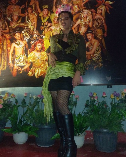 Me cosplaying as a gay cabbage. I'm in ruffles of various shades of green, and a headpiece made of red and pink flowers sit on top of my head. Strings of gold chains cascade from my head and frame my face.