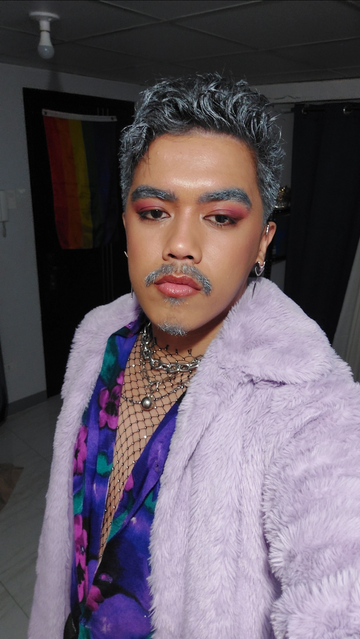 A selfie of myself dressed in an eye-catching outfit, featuring gray-streaked hair and detailed makeup. I am wearing a fluffy, lavender coat over a vibrant purple floral shirt, with a studded fishnet undershirt, a thick silver chain necklace, pink-hued eye shadow, and a light gray mustache and eyebrows.