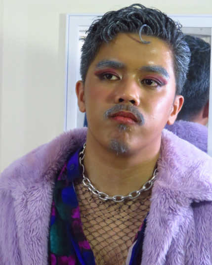 Gay dude wearing a lavender fur jacket over his purple floral top. His top is unbuttoned, revealing a studded mesh garment underneath and an oversized metal chain he is wearing around his neck. The guy's hair, eyebrows, mustache, and goatee are all colored silver gray. His eyelids are painted blush pink. He looks on to the distance. What is he up to?
