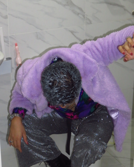 Guy with silver hair matching his glittery silver pants sitting on a covered toilet. He puts his left hand to a wall to support his weak and nauseous body. He bows his head down from being sick of the drinks he had. His scalp can be seen, revealing that his silver hair is not who he really is; it's all for show.
