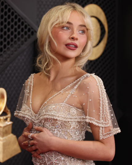 Sabrina Carpenter glancing off-camera while wearing a delicate, vintage-inspired cream gown intricately detailed with pearls and sequins. Her blonde hair is styled in soft waves with curtain bangs, complementing her classic red lip and the sheer, beaded sleeves of her dress.