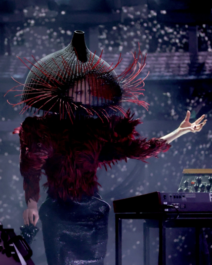 Lady Gaga performing her hit song "Abracadabra" at the 2026 Grammys while standing within a circle of synthesizers and keyboards. She wears an avant-garde cage-like headdress and a red-and-black feathered ensemble that creates a sharp, structured silhouette.