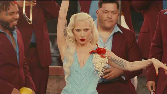 Lady Gaga in pale blonde hair, powder blue (almost light teal) dress, and a Hibiscus flower with jasmine strings as brooch attached to her left shoulder.