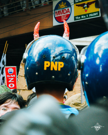 Taken during the protests on the 40th anniversary of the People Power Revolution in EDSA. A policeman wearing a helmet with PNP (Philippine National Police) obscures an effigy with horns, making it look like the policeman is growing the horns out of his own head.