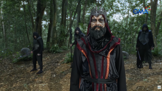 Hagorn standing in a forest wearing a dark fantasy costume made of lightweight, inexpensive-looking fabric, with a black robe trimmed in dull red material and a loosely structured bodice tied with a simple belt. He wears a rough, jagged crown and heavy face paint, while hooded figures in similarly plain black cloaks stand behind him holding weapons.