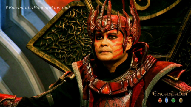 A regal-looking Hagorn sittinh on an ornate throne wearing a high-quality red and metallic armor ensemble with intricate sculpted detailing, layered plating, and a polished, jewel-accented horned helmet. The costume features rich textures, structured tailoring, and precisely applied face paint.