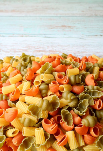 A vibrant pile of tricolor dry pasta, featuring shades of orange, green, and pale yellow. The pasta consists of various shapes, including ridged shells and spiral-like curls.
