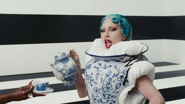 Lady Gaga dressed as a porcelain jar pouring tea out of her porcelain teapot into a porcelain teacup held by a disembodied hand.