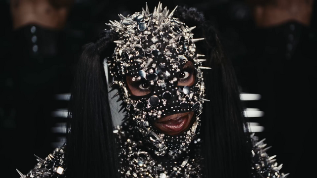 Doechii wearing a metal-studded headwear that covers her entire face, with openings for her eyes, mouth, and two black ponytails on each side of her head.