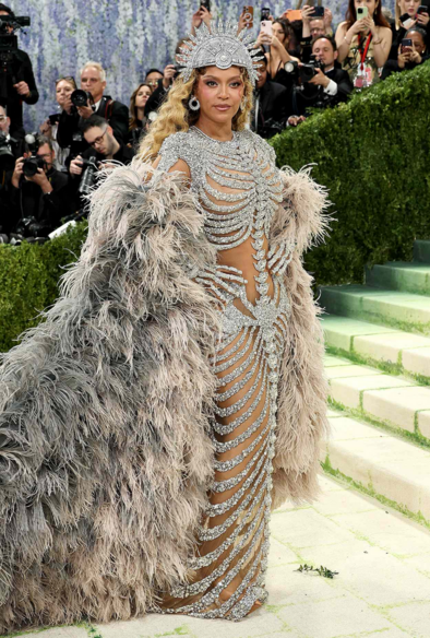 Beyoncé wearing a silver studded dress that looks like a skeleton. She also wears a long cape made of ostrich feathers and a studded cranial helmet headdress.
