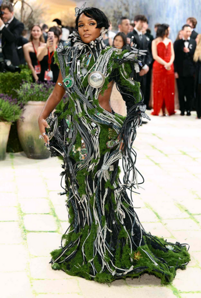 Jannelle Monaé in a mossy (literally) green dress covered in wires, computer ports, and connectors. She dons a butterfly hair clip on her head.