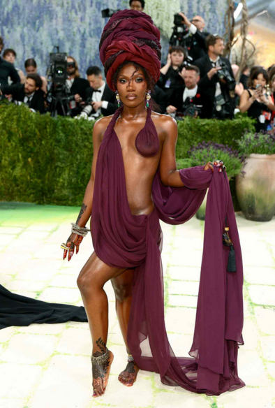 Doechii in a maroon Marc Jacobs outfit that drapes around her body and features an elaborate head wrap and bare feet, plus jewels including David Webb earrings and ring. 