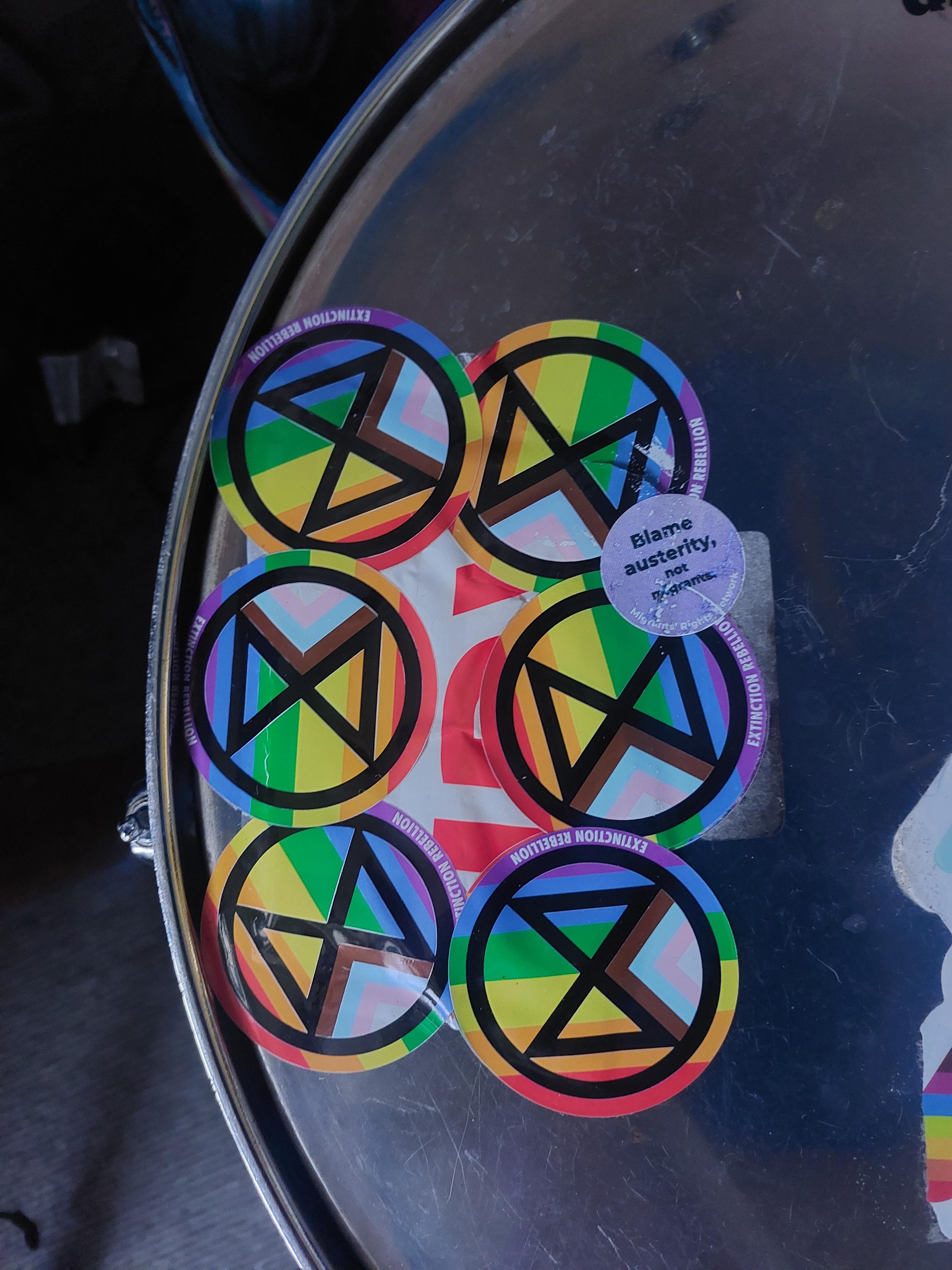 Edge of the skin on a DIY surdo (type of bass drum), showing the damping patch which is fundamentally duct tape but/and also fragile packing tape, a load of progressive pride XR stickers and 1 small purple "blame austerity, not refugees' sticker. Sounds great.