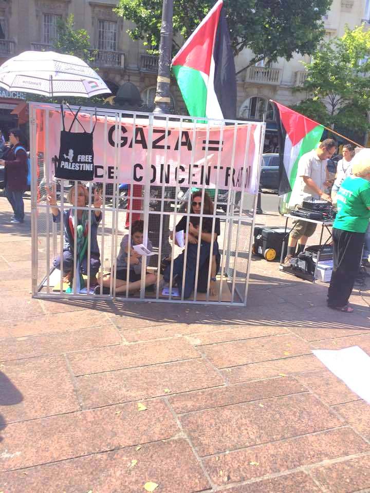 Manif pro-Palestine a l'exterieur, un mini-prison où il y a plusieurs personnes enfermées,  une affiche qui dit Gaza = Camp De Concentration. Plusieurs personnes autour avec des drapeaux palestiniens 

Photo donnée par @HarouatMed sur X