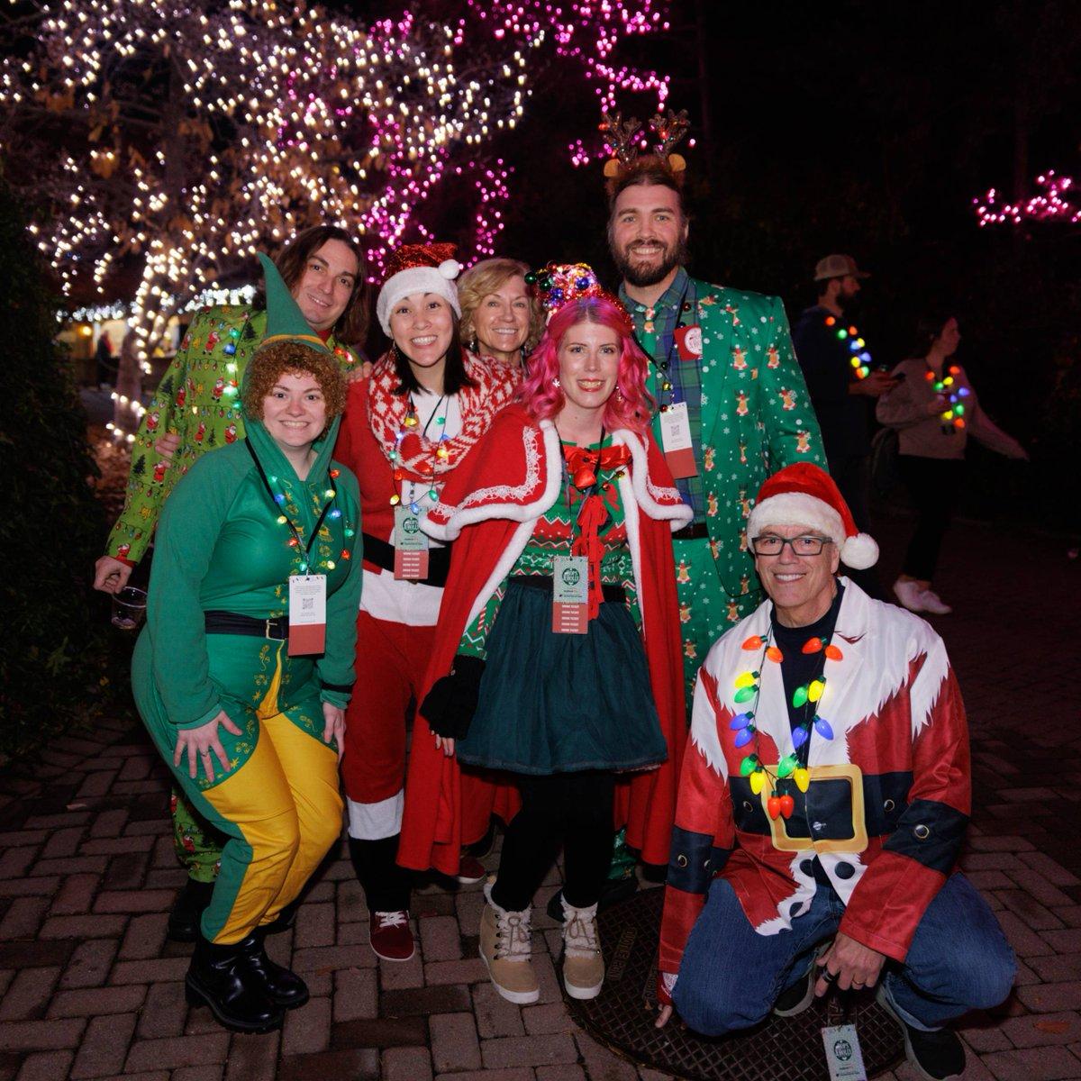 In this festive scene, a group of people dressed in costumes are gathered for a holiday celebration. There are seven individuals in total, all adorned with colorful attire, some wearing hats and others donning costumes that make them look festive and joyful. The costumes vary in design and color, with some people dressed in more elaborate outfits. The group appears to be posing for a picture, their smiles radiating the holiday spirit. In the background, there's a Christmas tree, further emphasizing the holiday theme of the photo.