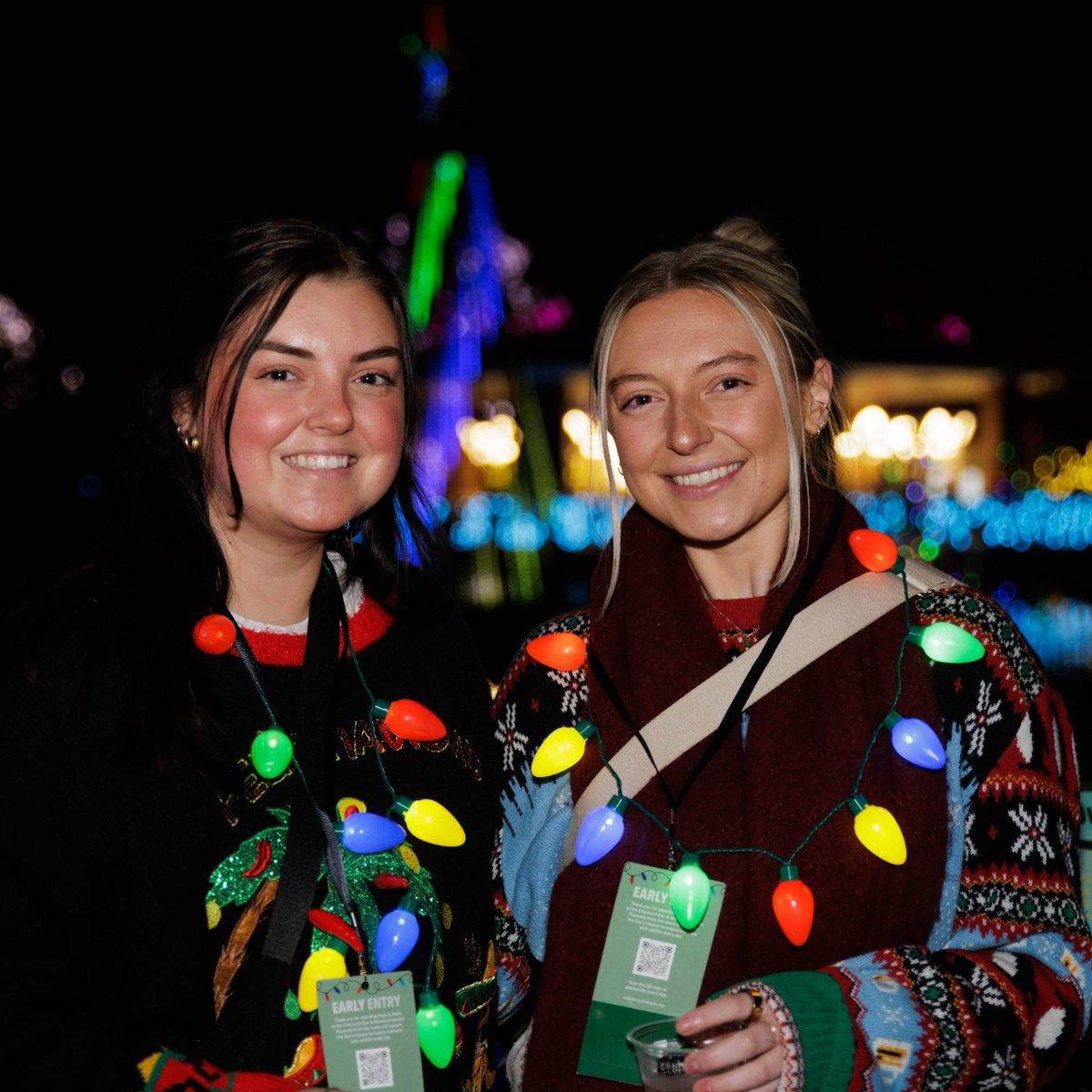 Two women in festive Christmas sweaters are standing side by side, smiling and holding drinks. One of the women is also wearing a necklace of multicolored Christmas lights. They are in front of a building with a tree, adding to the holiday atmosphere of the scene. The women appear to be enjoying each other's company, possibly having a casual celebration or outing during the holiday season.