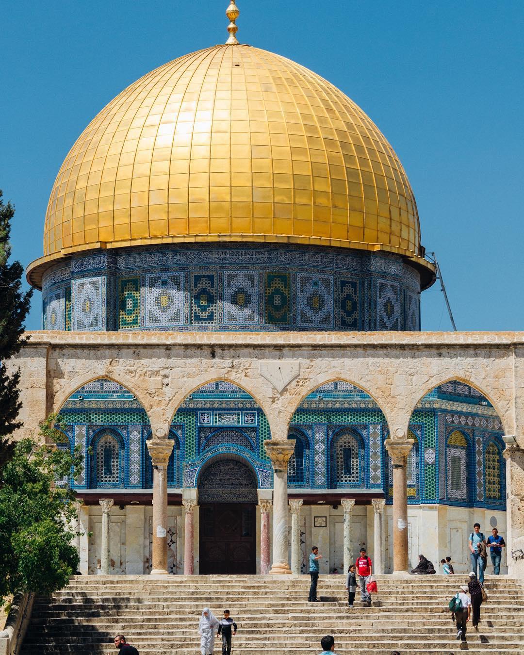 A low-angle shot of the Dome of the Rock in Jerusalem/Al Quds, featuring its iconic golden dome and intricate blue tilework, with people walking on the stone steps below. We also see the beige Al Mawaseen arches.