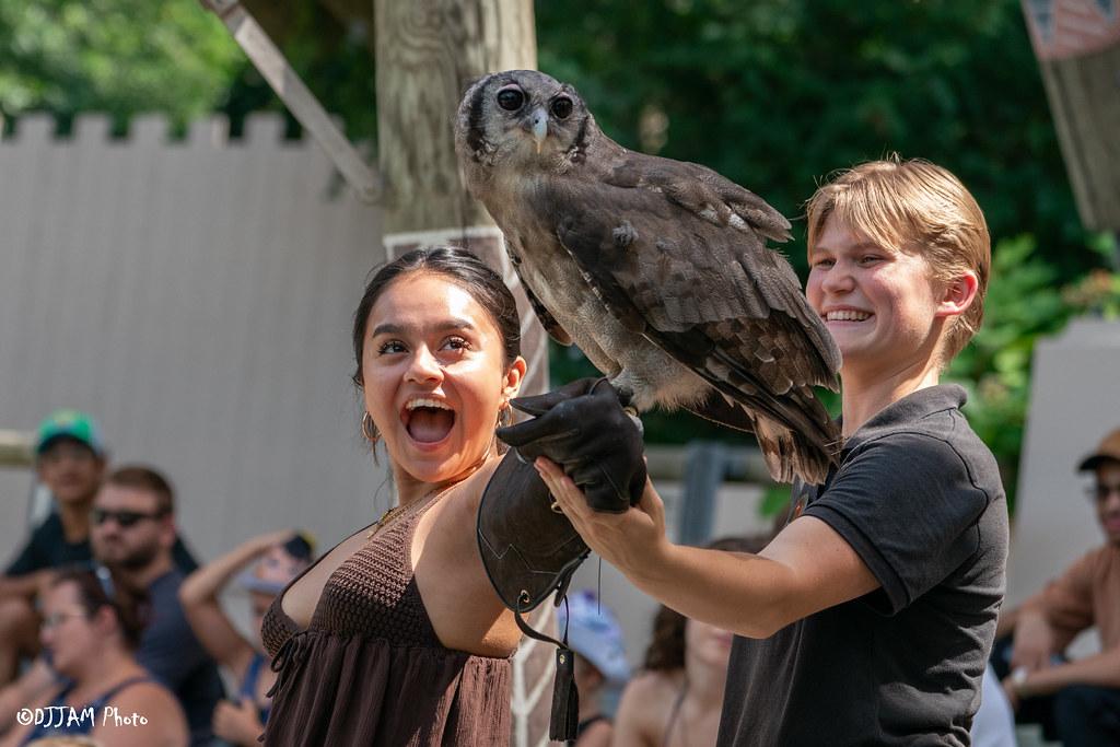 In the image, a man and a woman are standing together, holding a large bird on their shoulders. The woman is on the man's shoulders while the man is supporting the bird with his arms. They are both smiling and appear to be enjoying the moment, possibly as part of a bird show or event.

There are several other people in the scene, some standing behind the man and woman, observing their interaction with the bird. In the background, a potted plant can be seen, adding to the overall atmosphere of the location.
