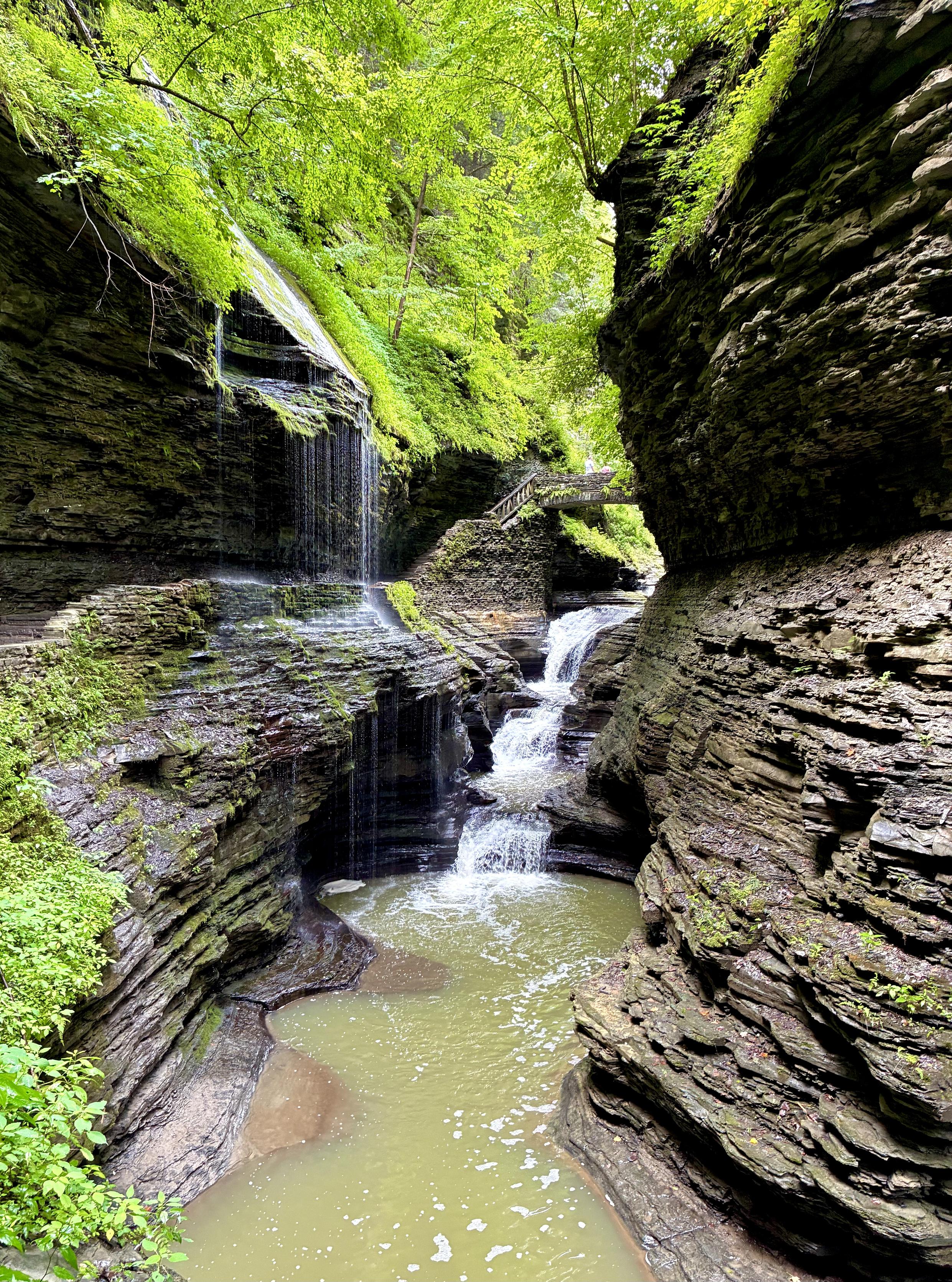 A narrow, winding gorge lies between towering layers of gray stone topped with green foliage. A stone trail runs along the left side, leading to a small stone bridge. Beneath the bridge, a small stream flows through, becoming a thin waterfall that cascades into a pool below. Another waterfall plunges from the top of the left cliff, falling like a veil or a curtain beside the stone trail.
