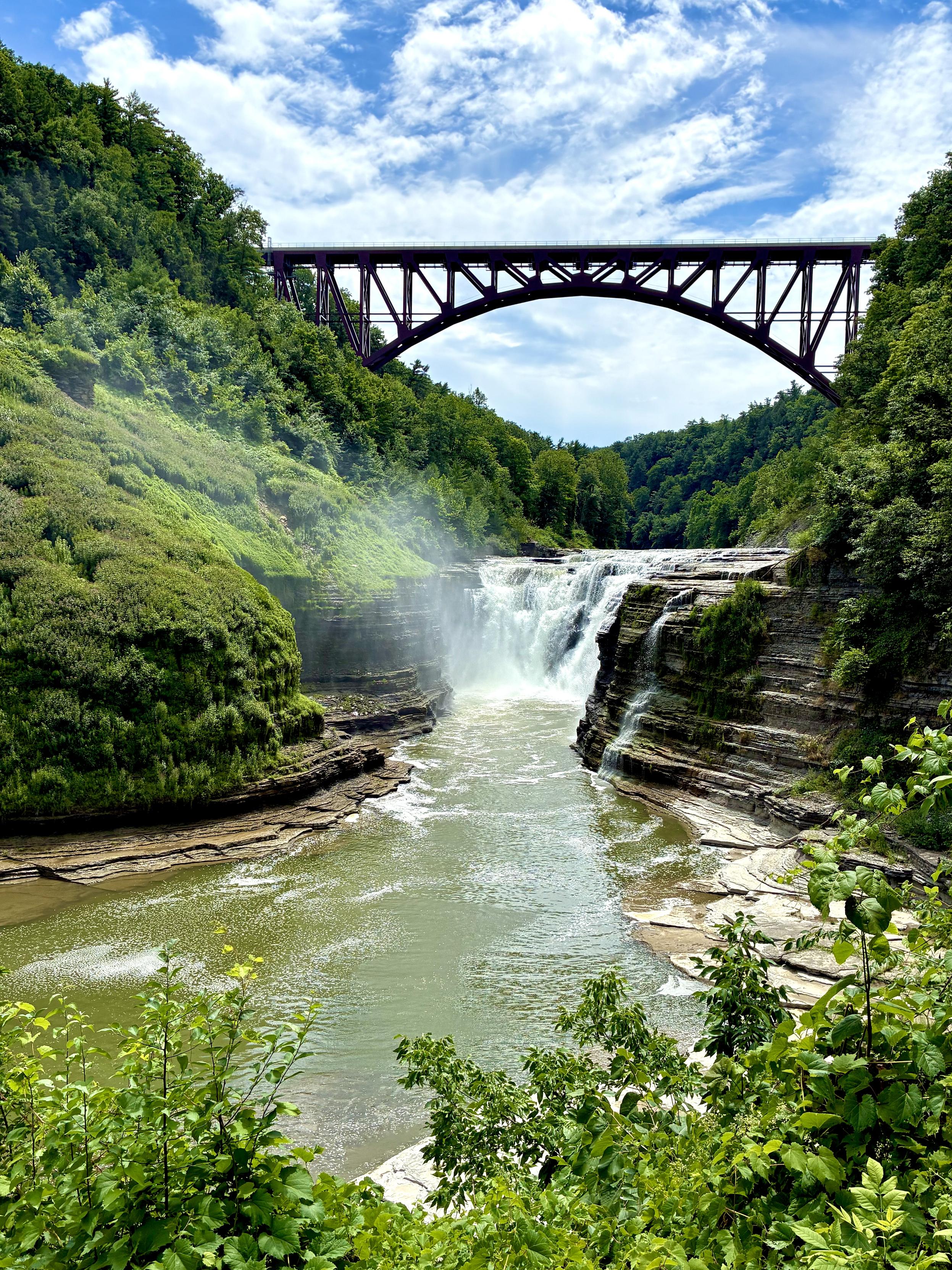 Under the partly sunny sky, river flowing in the valley plunged into the bottom of gorge flanked by the towering layers of gray stone bank on the both sides, with left banks covered by green. The plunged water spread out from the gorge bed and the murky swift water flows through the bottom. And a large red steel arch bridge spans the valley in the distance.