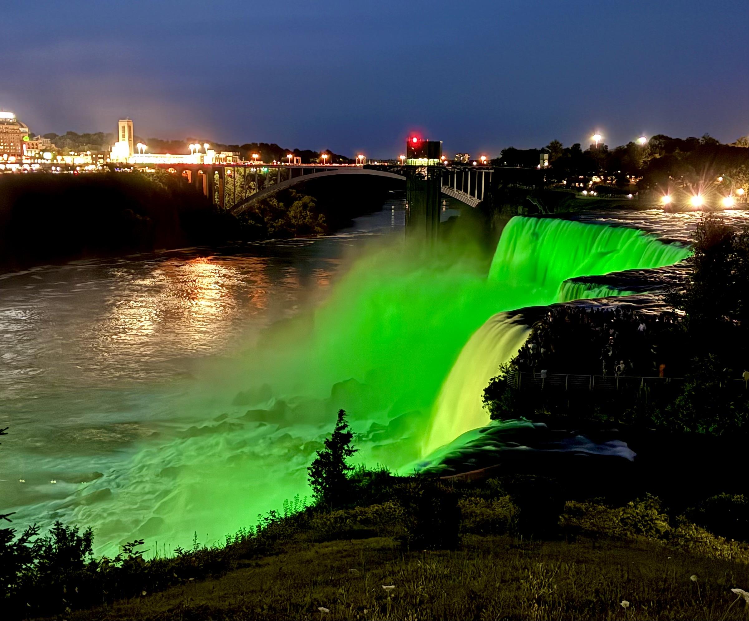 A nighttime view of American Falls from the edge of the cliffs, with the cascading water illuminated in vibrant green light. The top part of Bridal Vail Falls is also visible in the foreground.