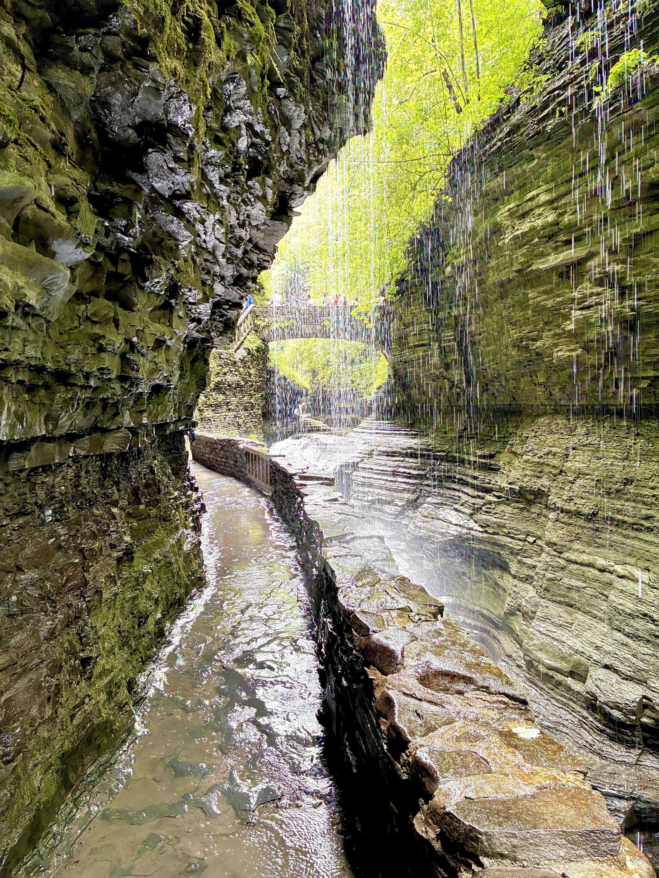 A stone path along the left side of the narrow gorge leads to a stone bridge in the distance. Towering walls of layered gray rock rise on both sides, while a curtain of cascading water flows over part of the path. The gray rock walls are softly tinted green, reflecting the vibrant forest visible through the narrow opening above the gorge.