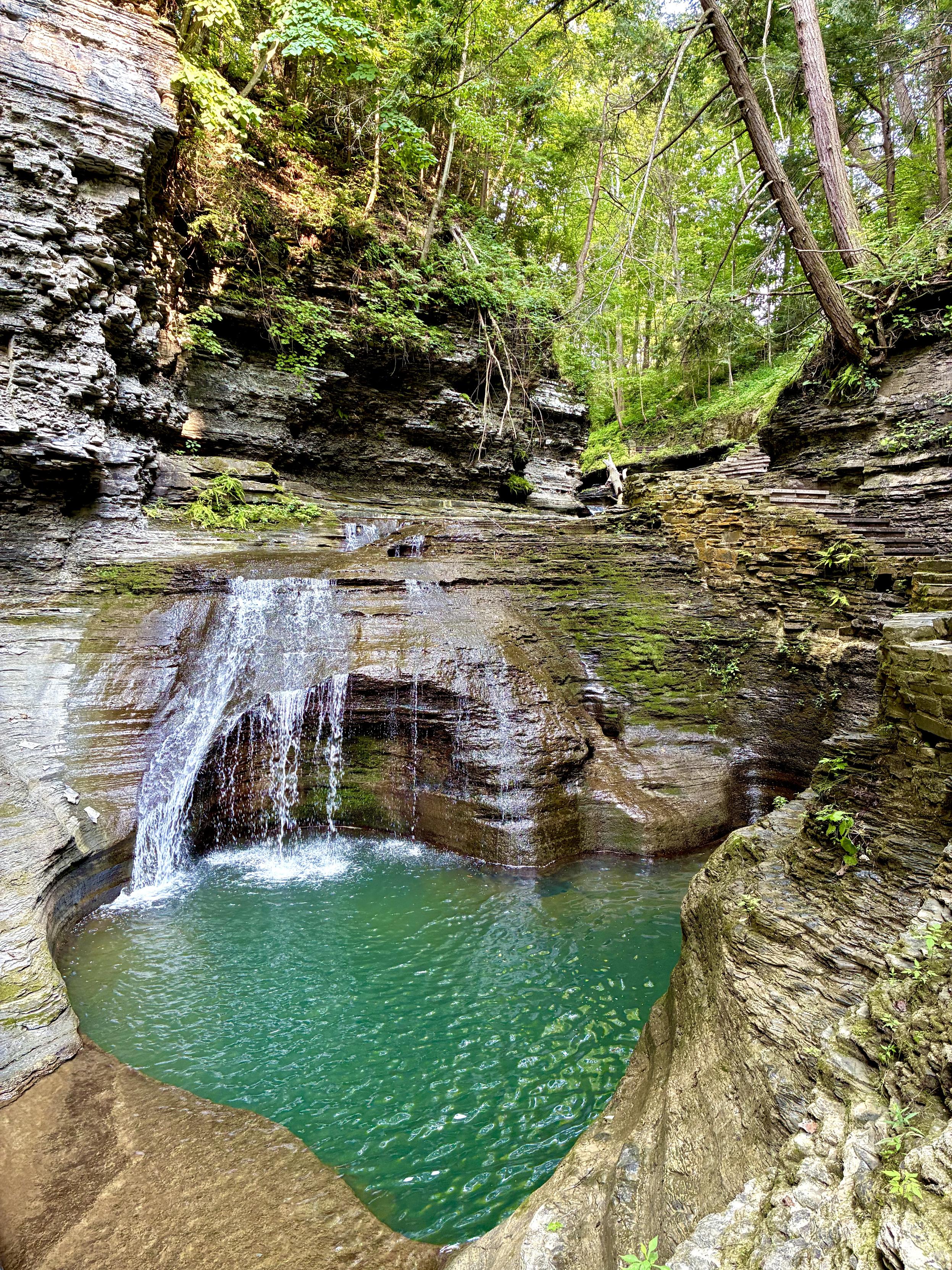 A small waterfall cascades over layered rock into a turquoise pool in a narrow, lush forest gorge. Steep, stratified rock walls rise on both sides, with a stone stairway winding along the right side beneath dense green trees.